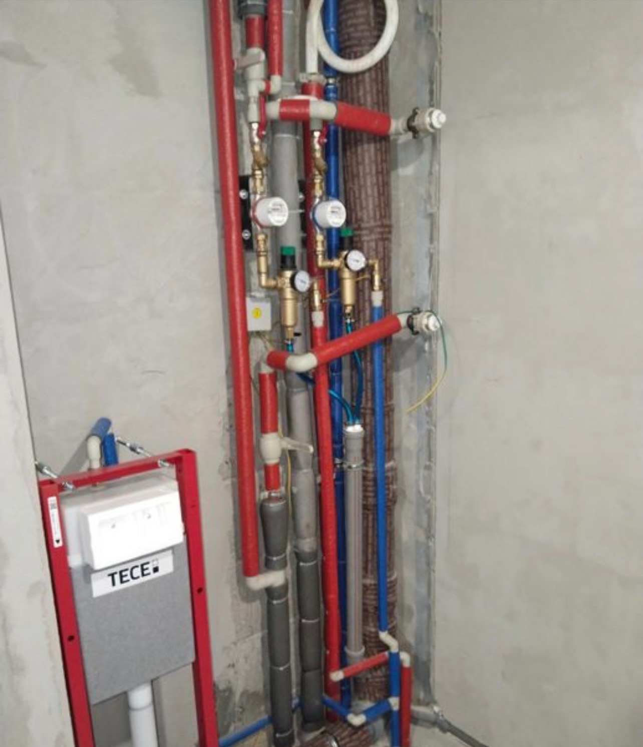 Instalacja wodno-kanalizacyjna w surowej łazience, widoczne rury w kolorach czerwonym i niebieskim, manometry oraz stelaż podtynkowy TECE.