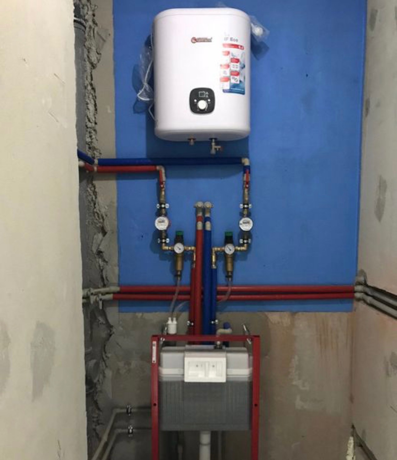 Bojler elektryczny nad stelażem WC na niebieskiej ścianie, z widoczną instalacją hydrauliczną z rurami w kolorach czerwonym i niebieskim. Ściany w trakcie wykończenia.