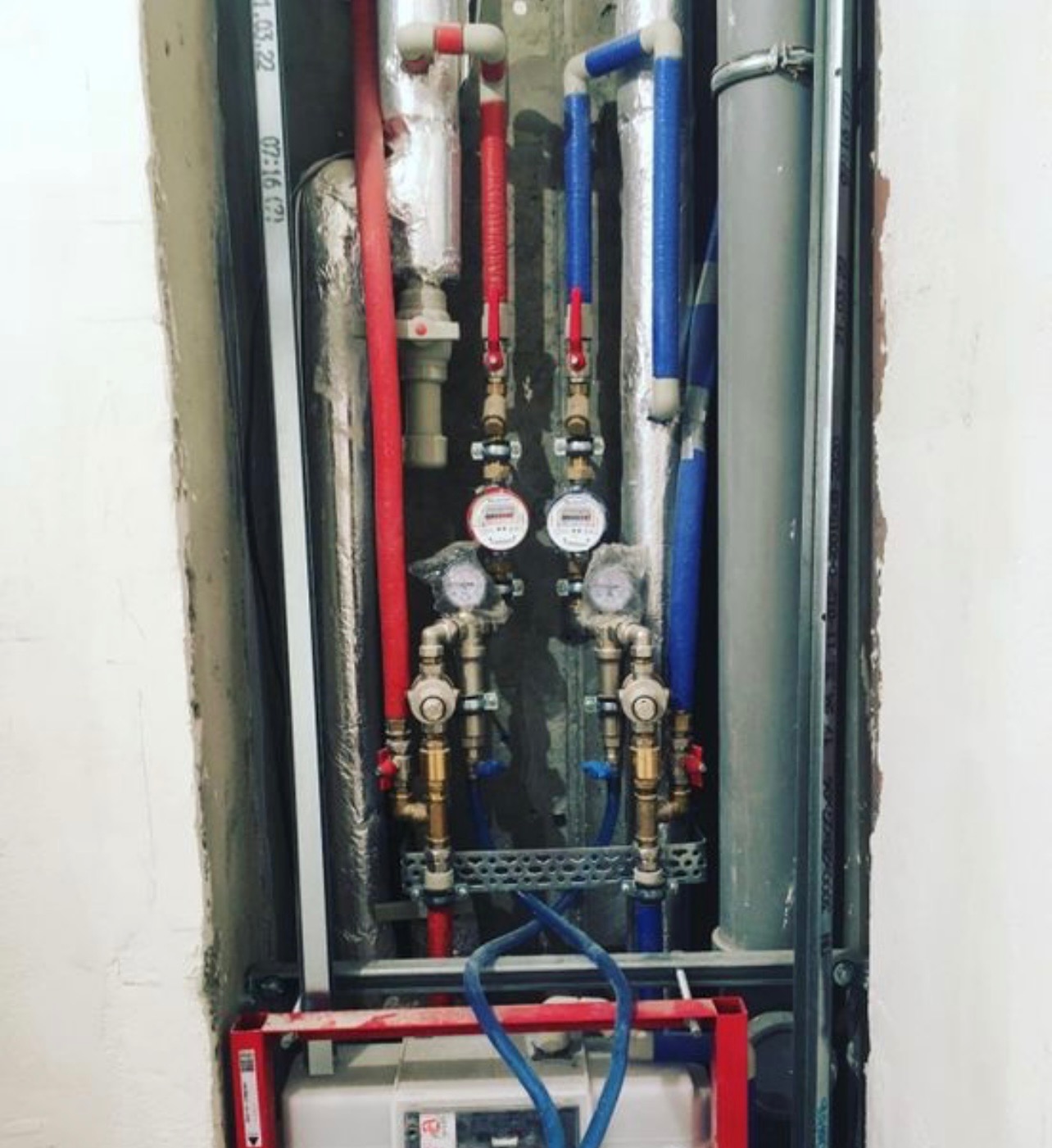 Instalacja wodna z rurami (czerwone, niebieskie) i licznikami w surowej łazience. Widoczne elementy montażowe i fragment zbiornika. Ujęcie z bliska, detale instalacji.