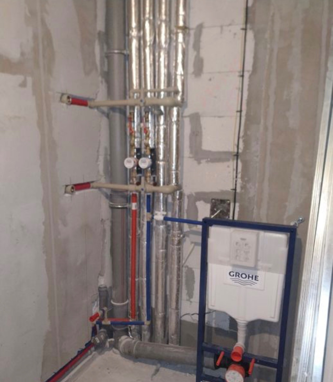 Surowa ściana z widoczną instalacją hydrauliczną w srebrnej izolacji, rurami w kolorze szarym, niebieskim i czerwonym oraz stelażem podtynkowym Grohe w łazience w trakcie remontu.