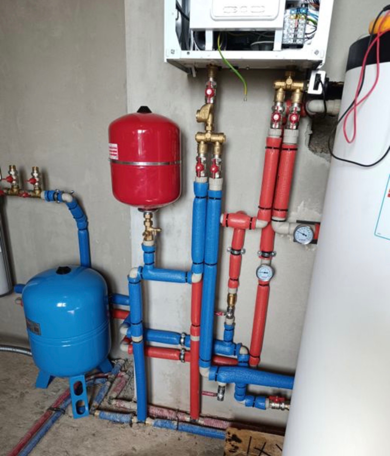 Instalacja CO z czerwonym zbiornikiem wyrównawczym i niebieskim naczyniem przeponowym, widoczne rury w izolacji, manometry i zawory. Szara ściana w tle.