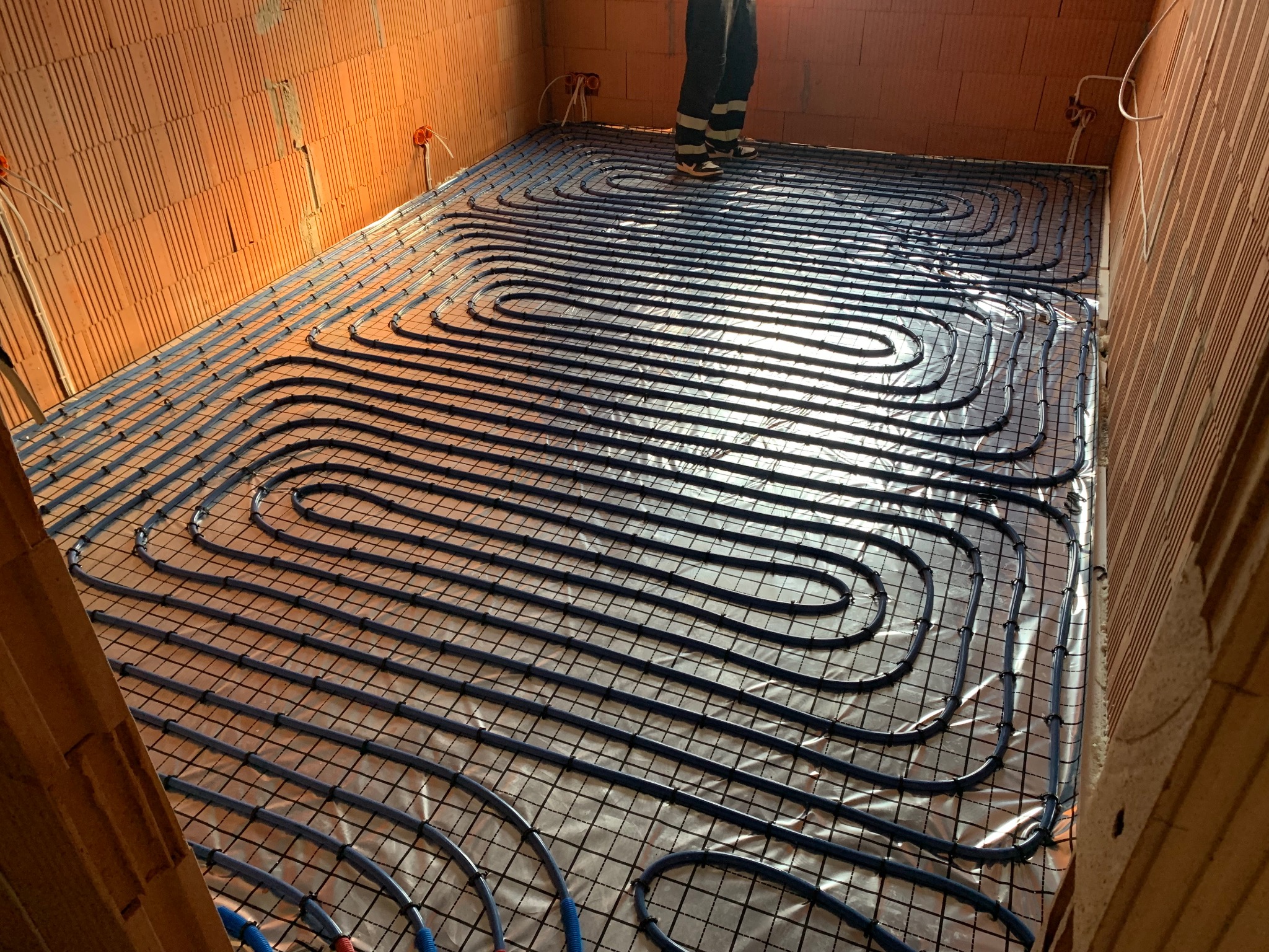 Ułożone, niebieskie rury ogrzewania podłogowego na folii, widoczne fragmenty ścian z cegły dziurawki i instalacja elektryczna. Częściowo widoczna osoba w roboczym ubraniu.