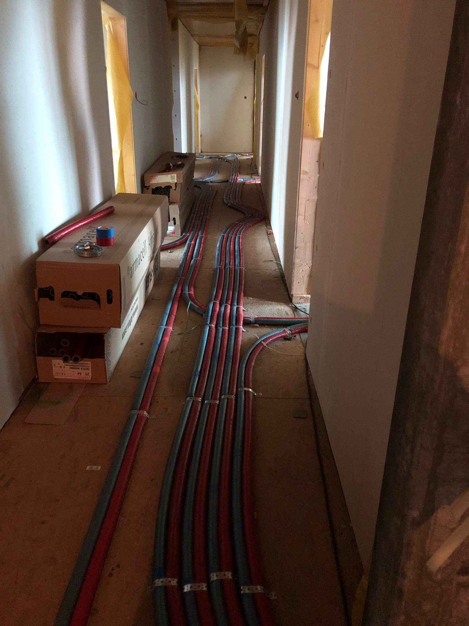Instalacja grzewcza podłogowa w korytarzu w trakcie montażu. Widoczne rury w kolorze czerwonym i niebieskim, prowadzone wzdłuż ścian. Na podłodze kartony z materiałami.