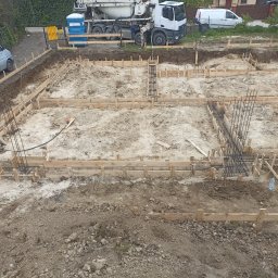 Marcin Chodyna - Przygotowanie terenu pod fundamenty domu: widok z góry na wykop z drewnianym szalunkiem i zbrojeniami, w tle betoniarka i samochody dostawcze.