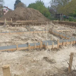 Wylany beton w szalunkach fundamentowych, wystające pręty zbrojeniowe, pryzma ziemi w tle.