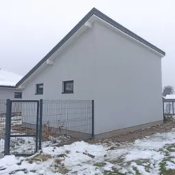 Nowoczesny, szary dom jednorodzinny z dachem dwuspadowym i czarnymi oknami, ogrodzony metalową siatką, częściowo ośnieżony.