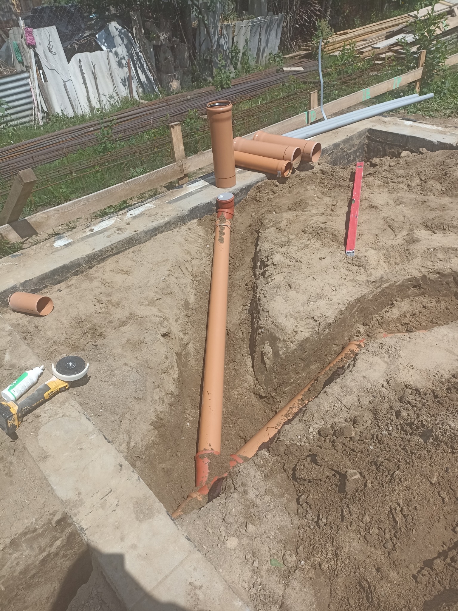 Wykop pod fundament z ułożonymi rurami kanalizacyjnymi, widoczny poziomica, szlifierka kątowa i klej. Na obrzeżach widać zbrojenie.