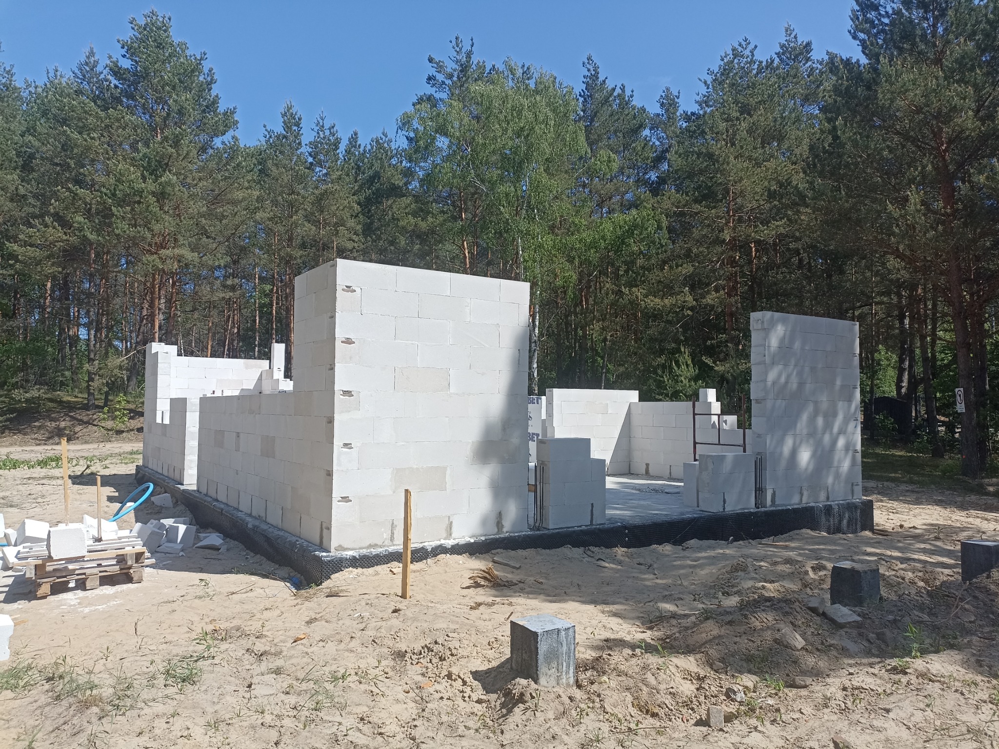 Stan surowy otwarty domu jednorodzinnego z betonu komórkowego w Długosiodle, widoczne fundamenty i pierwsze ściany na tle lasu.