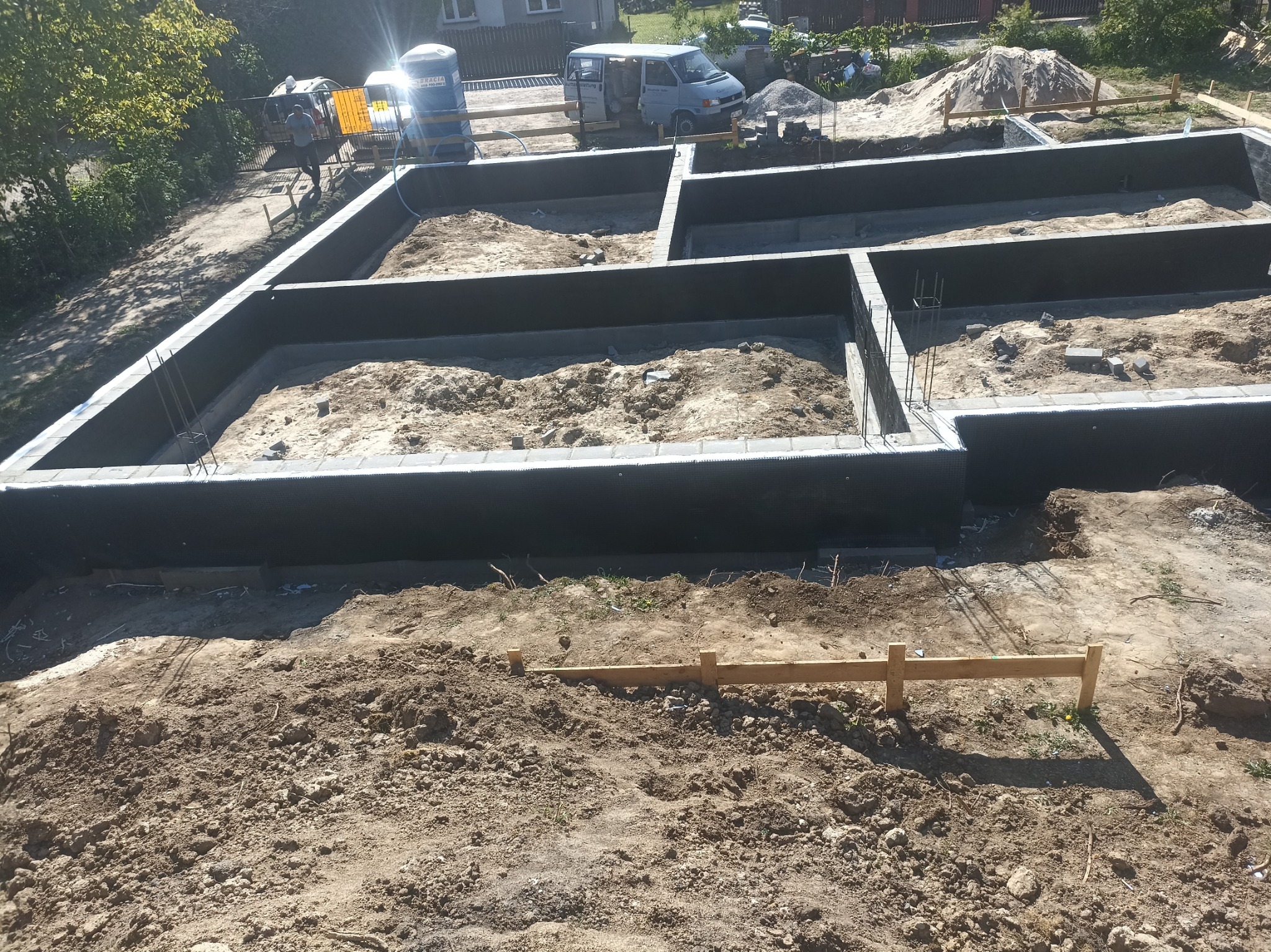 Zalane fundamenty domu jednorodzinnego z widocznymi prętami zbrojeniowymi, deskowaniem i przygotowanym terenem pod budowę.