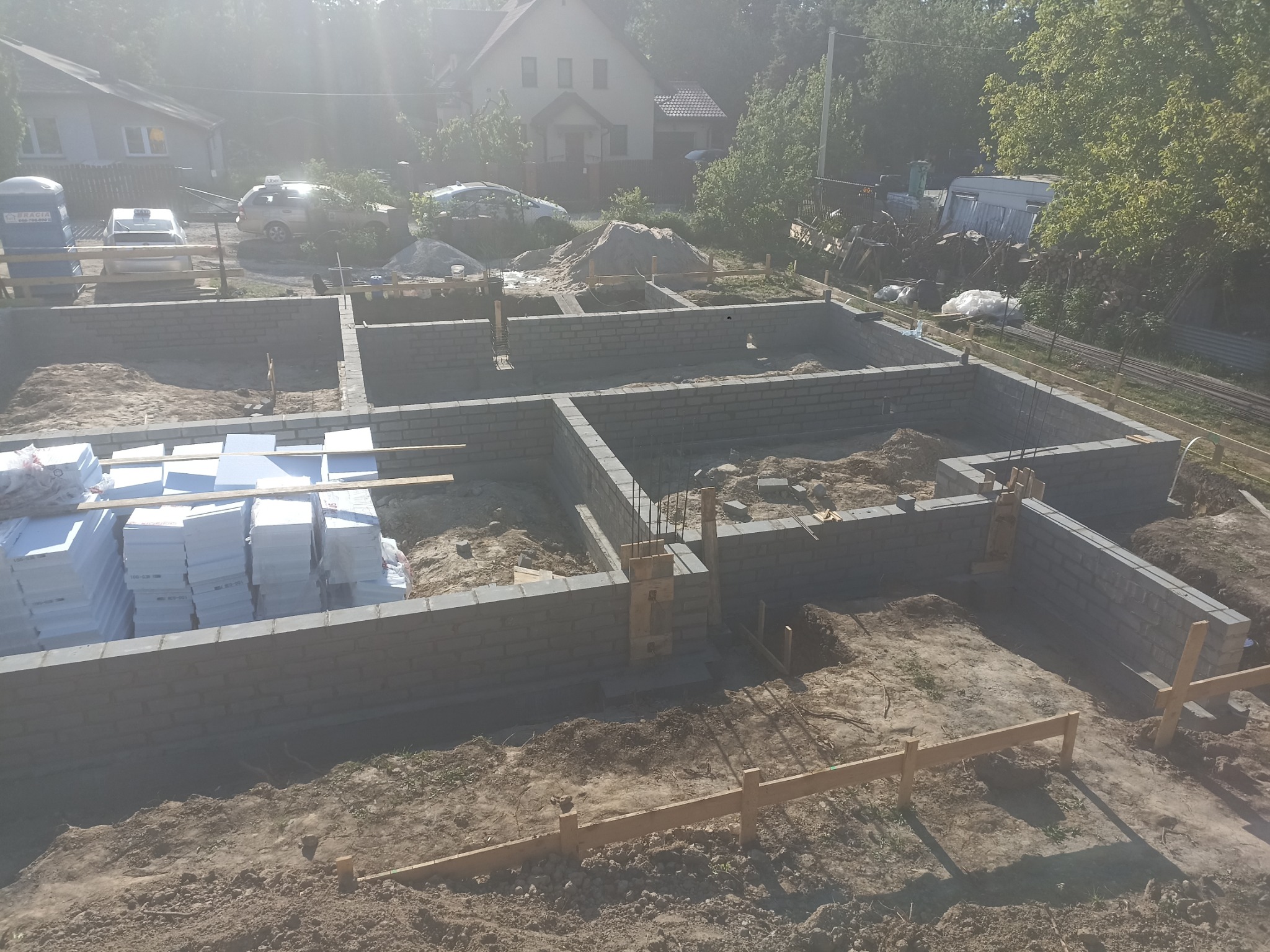 Wznoszenie fundamentów z bloczków betonowych; widoczne zbrojenia, drewniane szalunki i stos białych płyt izolacyjnych na placu budowy.