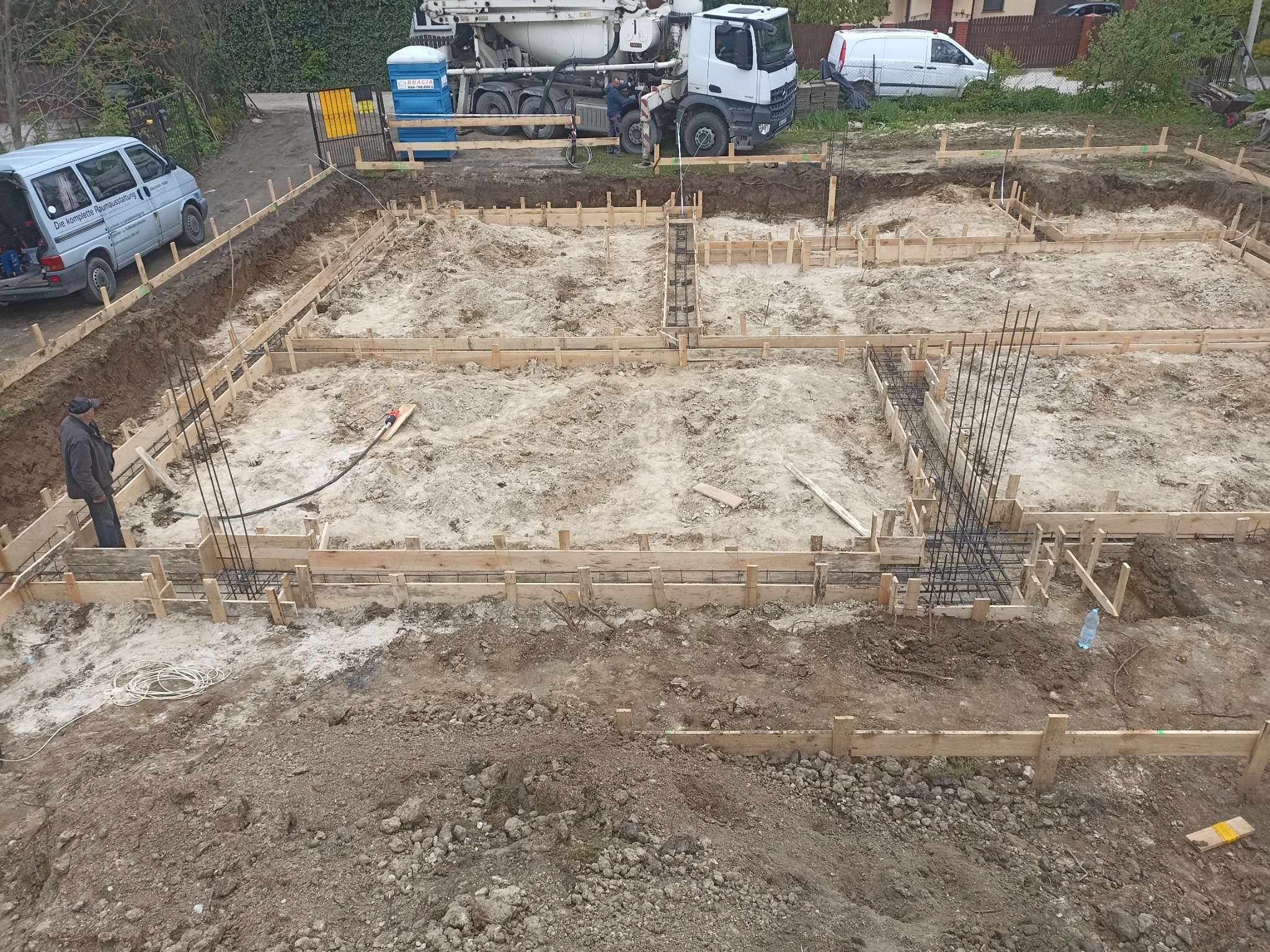 Przygotowanie terenu pod fundamenty domu: widok z góry na wykop z drewnianym szalunkiem i zbrojeniami, w tle betoniarka i samochody dostawcze.