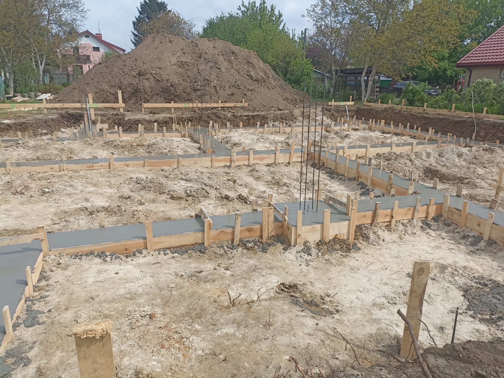 Wylany beton w szalunkach fundamentowych, wystające pręty zbrojeniowe, pryzma ziemi w tle.