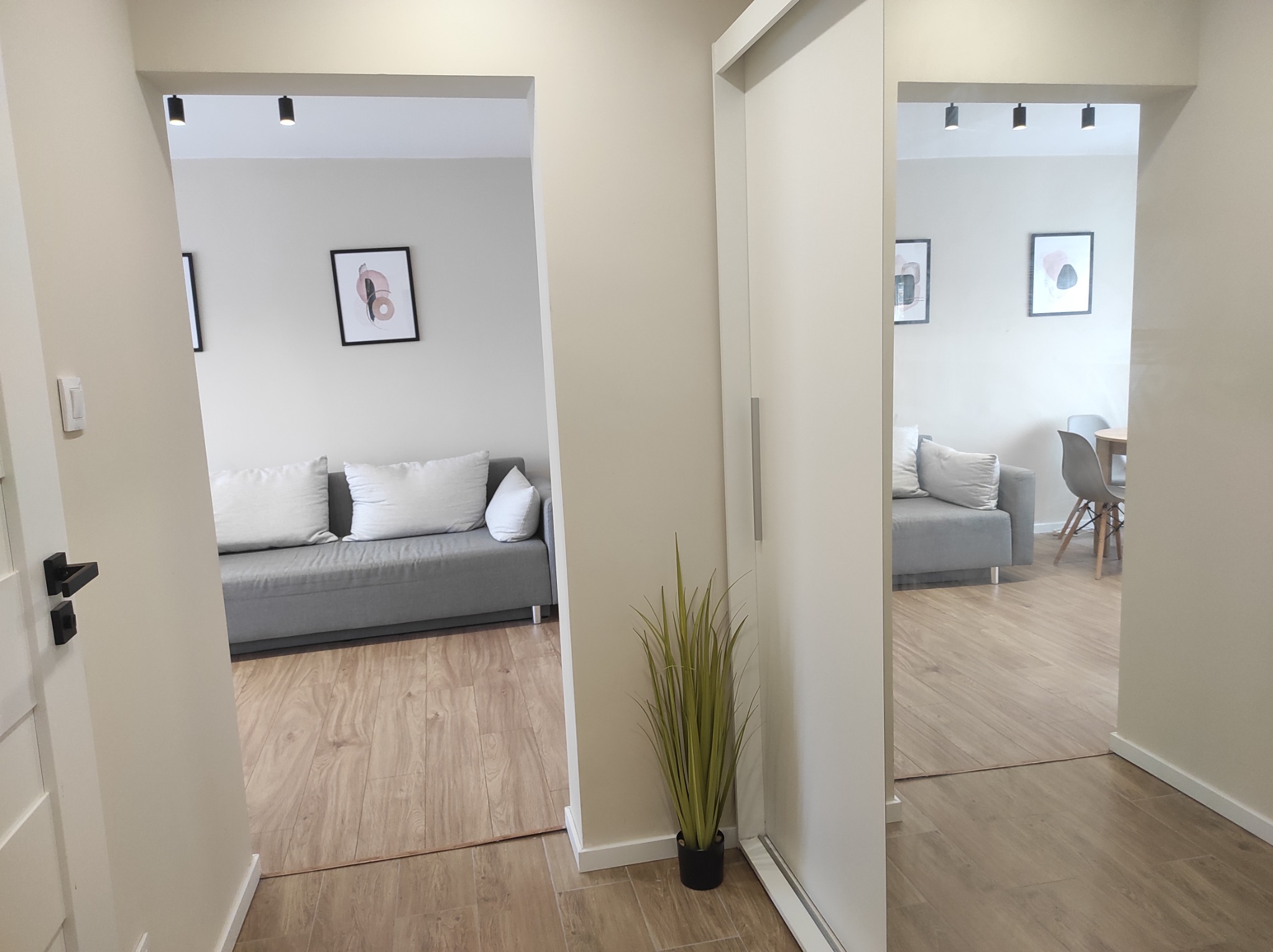 Widok z korytarza na dwa pokoje, w jednym szara sofa z białymi poduszkami i abstrakcyjne obrazy na ścianie, w drugim widoczny stół z krzesłami; podłoga wyłożona panelami imitującymi drewno,...
