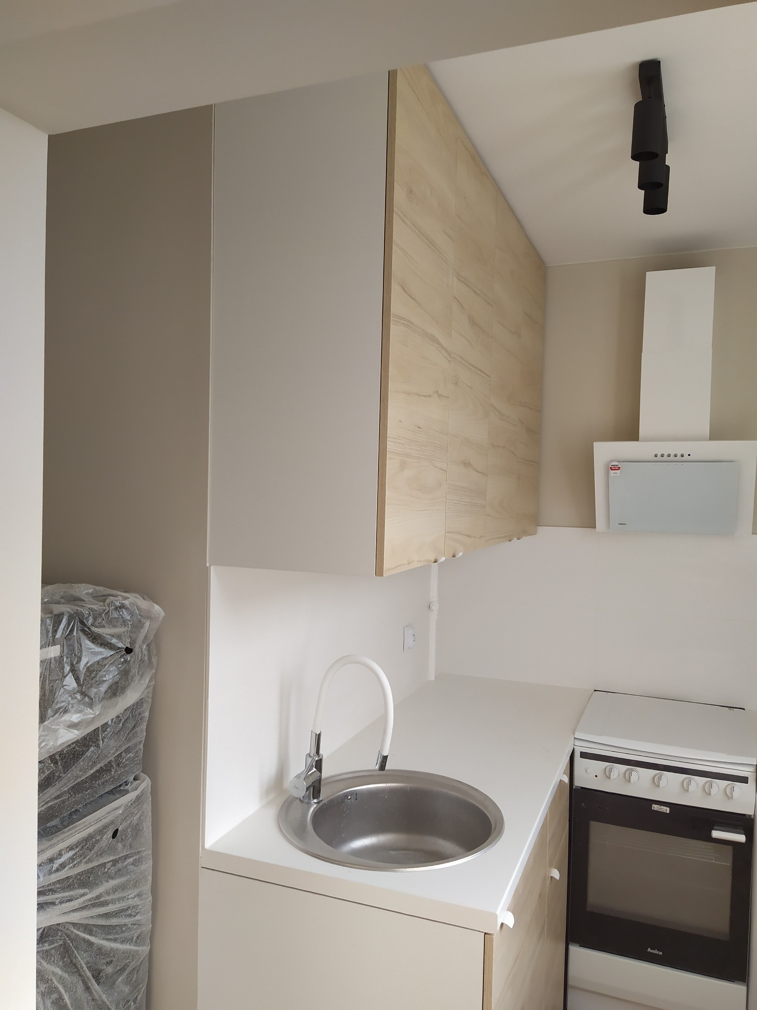 Jasna kuchnia z drewnianymi akcentami: okrągły zlew, biały blat, piekarnik Amica, szafki wiszące z frontami imitującymi drewno, okap i czarna lampa sufitowa. Po lewej widoczne elementy zapakowane...