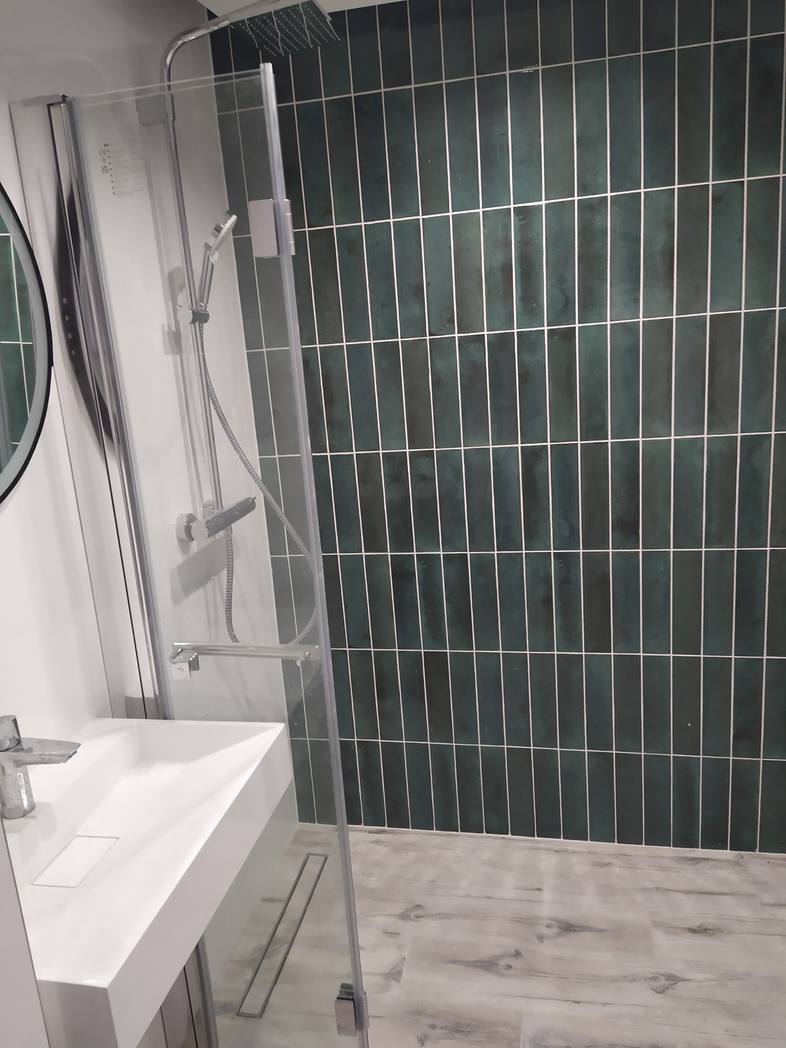 Nowoczesna łazienka z kabiną prysznicową ze szklanymi drzwiami, umywalką nablatową i pionowymi, zielonymi płytkami na ścianie.