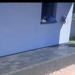 Ułożona, ciemnoszara kostka brukowa przed budynkiem o niebieskiej elewacji, z widocznym oknem, na którym leżą przedmioty, oraz gniazdem elektrycznym na ścianie.