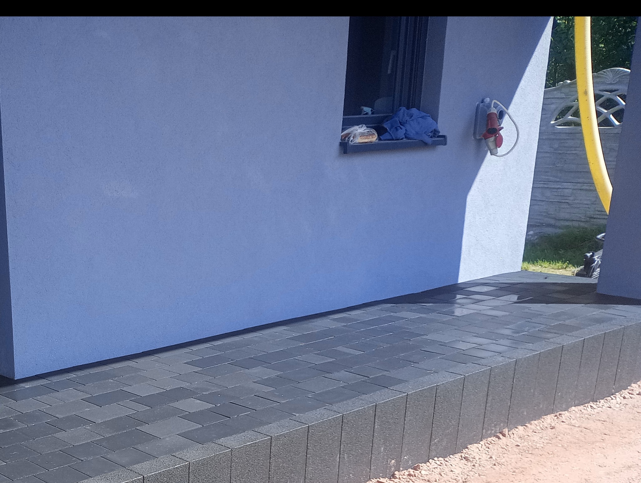 Ułożona, ciemnoszara kostka brukowa przed budynkiem o niebieskiej elewacji, z widocznym oknem, na którym leżą przedmioty, oraz gniazdem elektrycznym na ścianie.