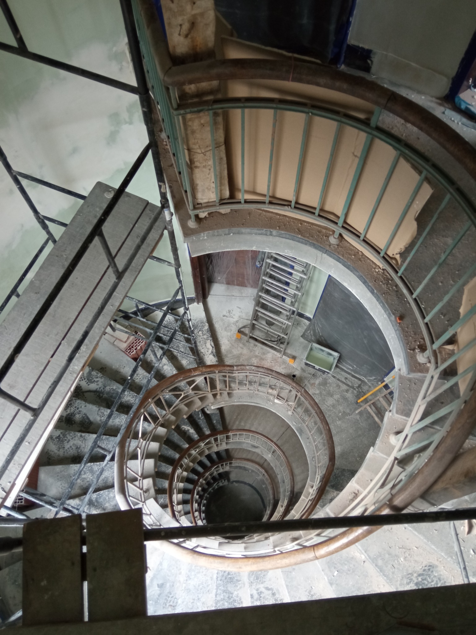 Wysoki kąt na spiralne schody w remoncie, z widocznymi drabinkami, folią ochronną, wiadrem z farbą i częściowo odrapanymi ścianami.