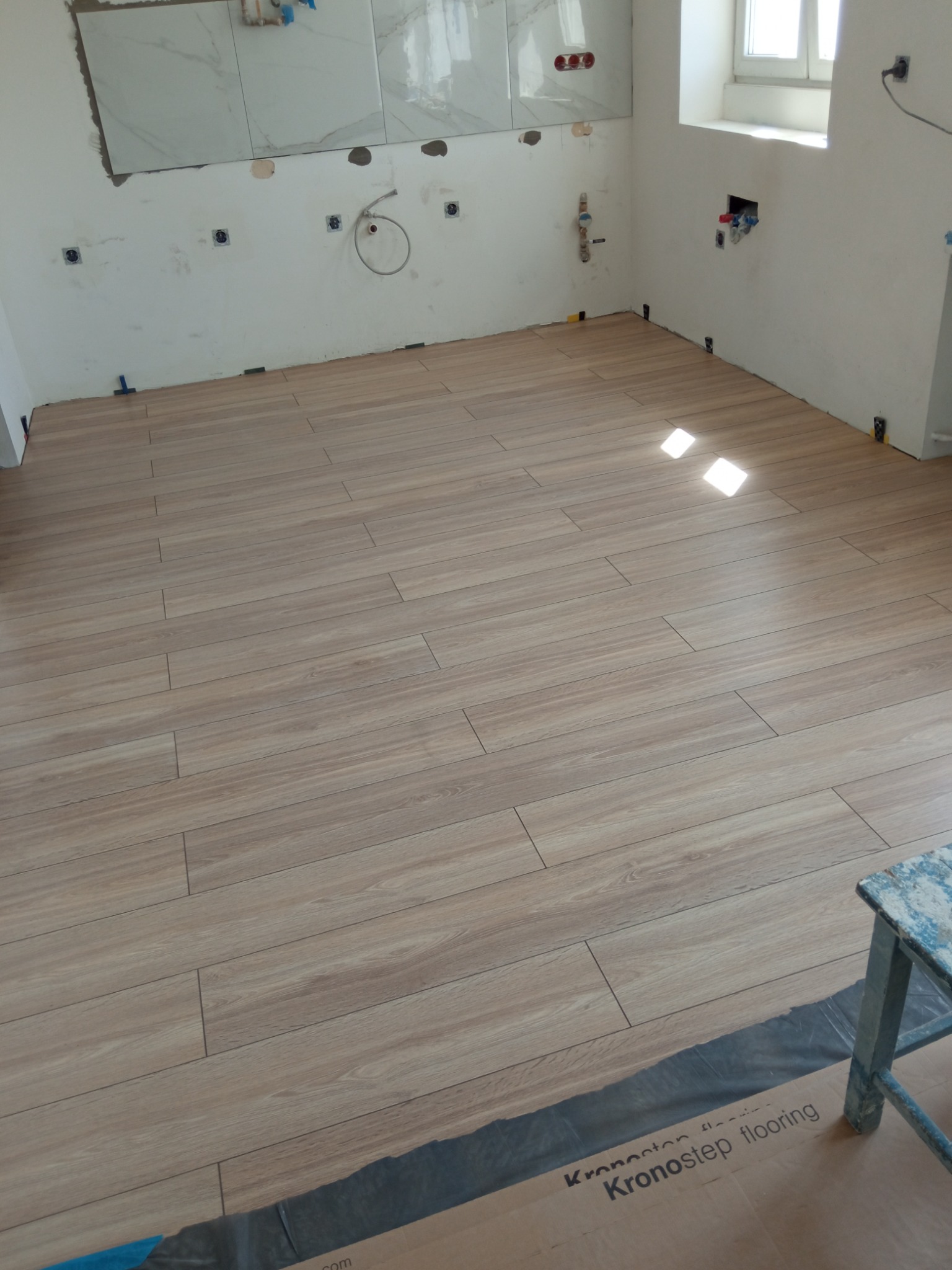 Ułożona podłoga z paneli imitujących drewno w trakcie remontu łazienki, widoczne przygotowane ściany pod płytki i instalacje, oraz resztki opakowania paneli podłogowych Kronostep flooring.