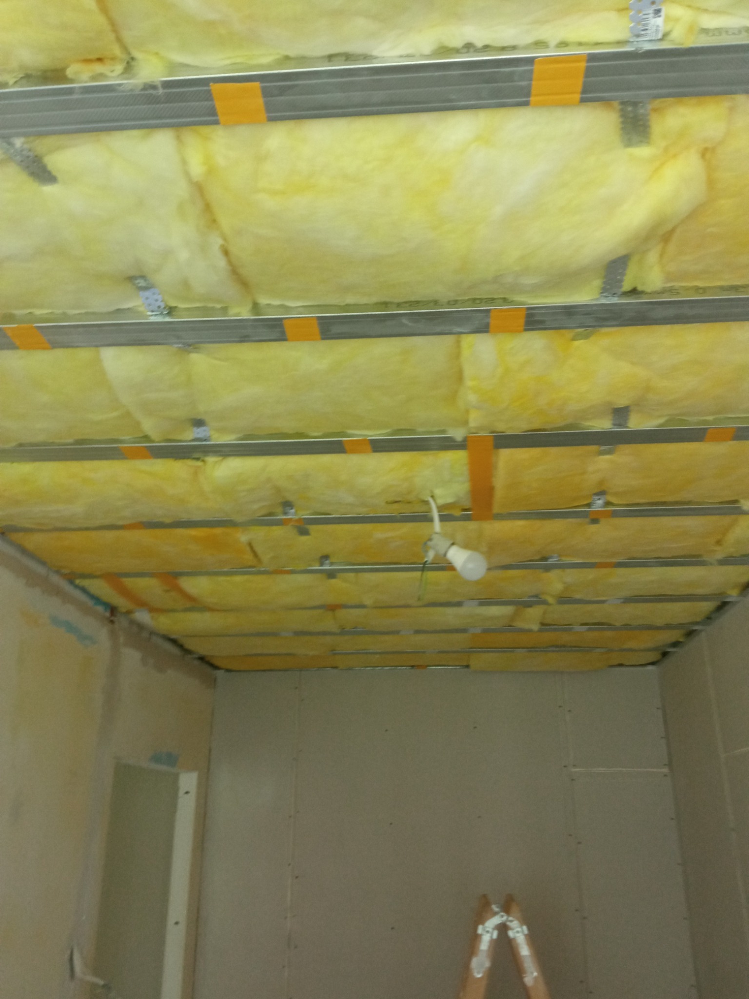 Montaż izolacji z wełny mineralnej w suficie podwieszanym, widoczne profile metalowe, taśma klejąca i tymczasowe oświetlenie w pomieszczeniu w trakcie prac wykończeniowych.