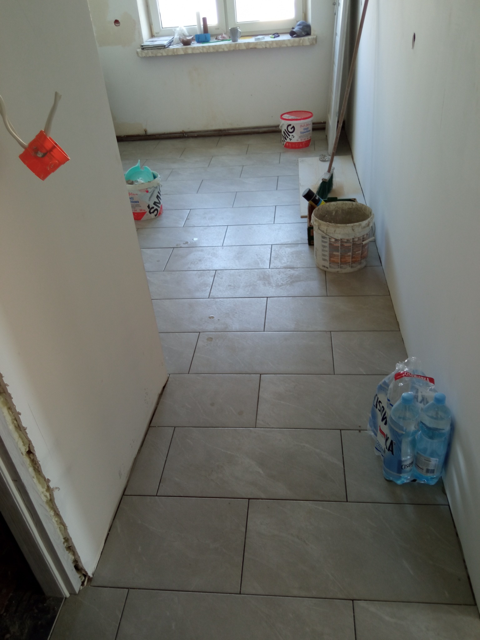 Ułożona podłoga z szarych, prostokątnych płytek w trakcie prac wykończeniowych w łazience, widoczne wiadro z zaprawą, butelki wody mineralnej i puszka z farbą.