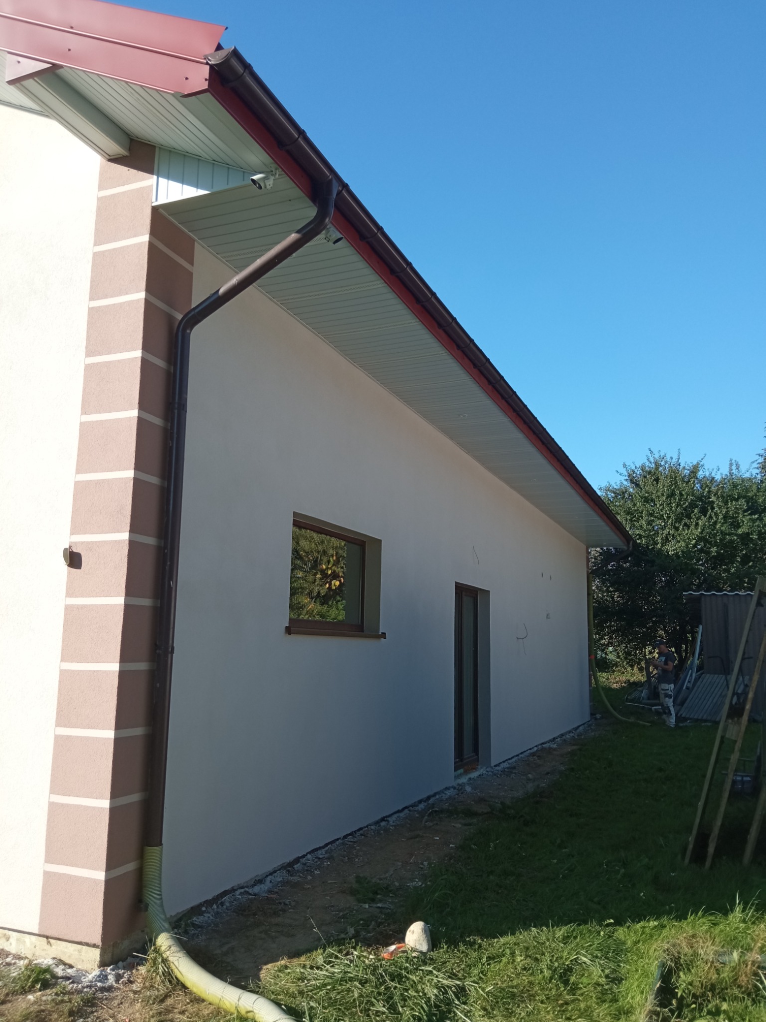 Świeżo otynkowana elewacja domu jednorodzinnego z brązowymi akcentami na narożniku i wokół okna, widoczna rynna i fragment dachu, w tle zieleń drzew.