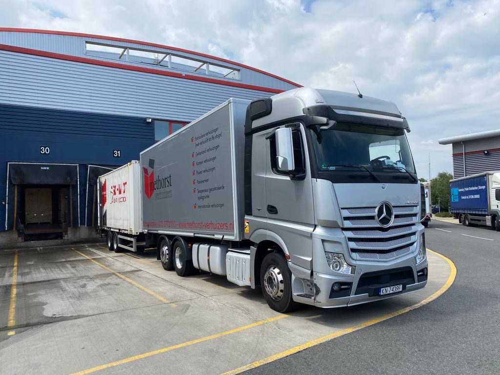 Srebrna ciężarówka Mercedes z naczepą kontenerową Methorst, zaparkowana przed magazynem z numerami 30 i 31, widoczne napisy na naczepie w języku niderlandzkim.
