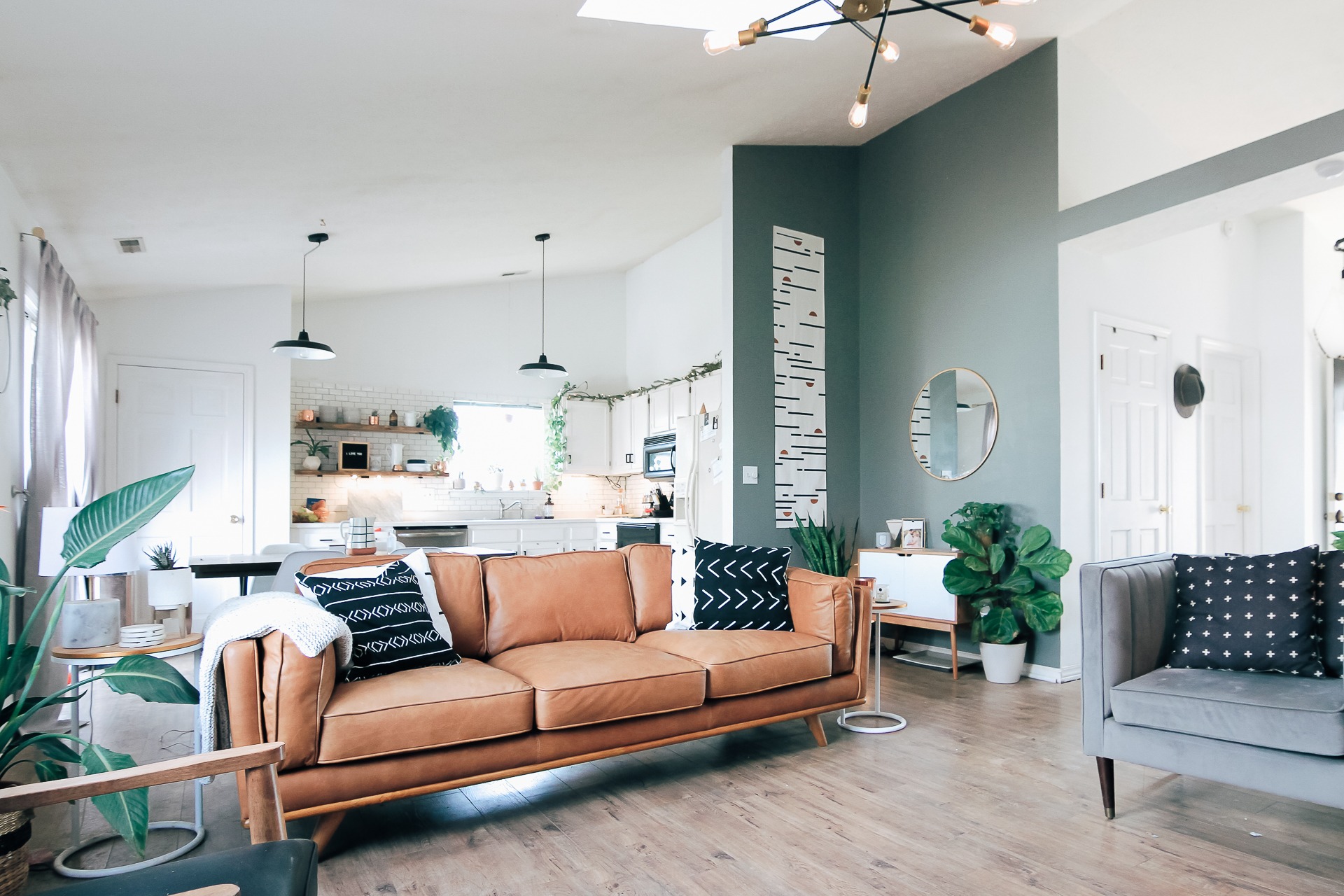 Jasne, przestronne wnętrze z brązową skórzaną sofą, szarą ścianą akcentującą z okrągłym lustrem, drewnianą podłogą i otwartą kuchnią w tle.