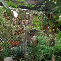 Ogród z drewnianą pergolą i dekoracjami, w tle krata i roślinność. Elementy ceramiczne i metalowe wiszą na konstrukcji, tworząc unikalny wystrój.