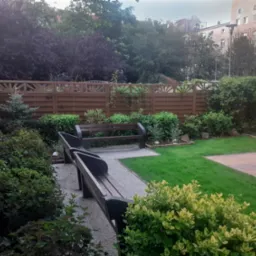 Uporządkowany ogród z dwiema ławkami, trawnikiem i ozdobnymi krzewami. Drewniany płot z ażurowym wykończeniem w tle. Elementy małej architektury ogrodowej.