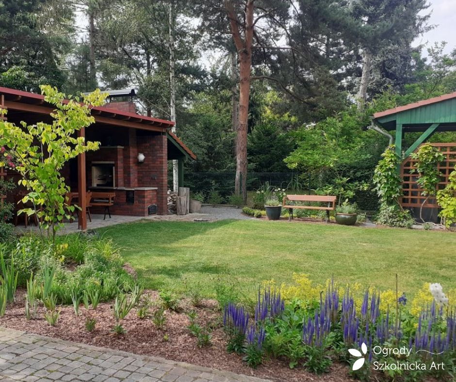 Przestronny ogród z zadbanym trawnikiem, rabatami kwiatowymi z niebieskimi i żółtymi kwiatami, murowaną kuchnią letnią z grillem, drewnianą ławką i altaną w tle.