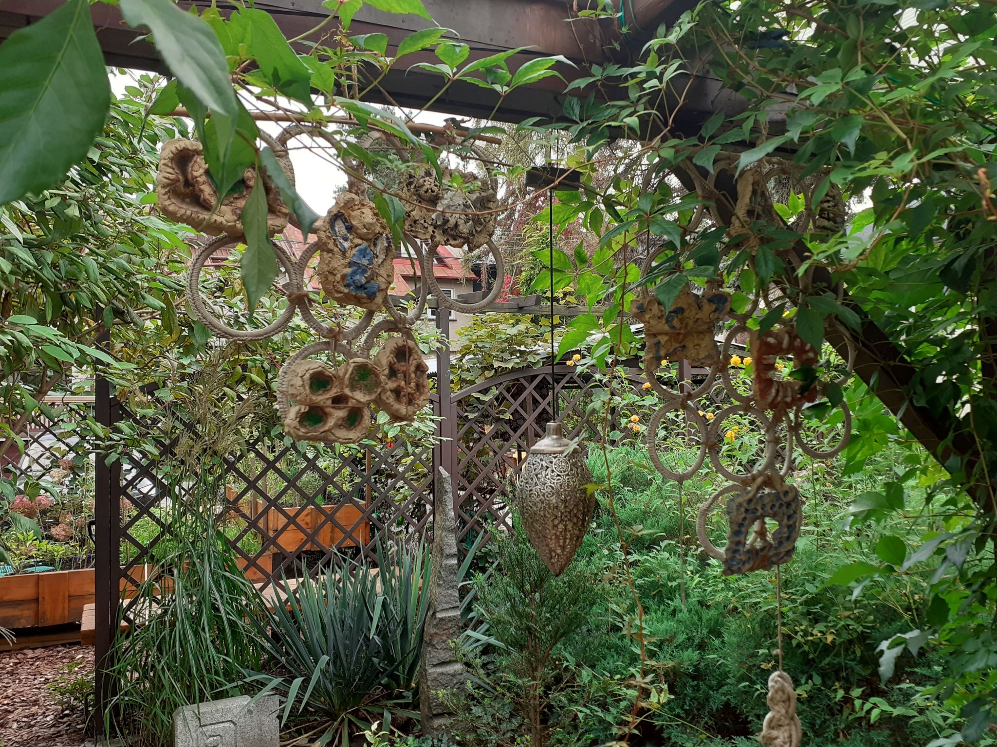 Ogród z drewnianą pergolą i dekoracjami, w tle krata i roślinność. Elementy ceramiczne i metalowe wiszą na konstrukcji, tworząc unikalny wystrój.