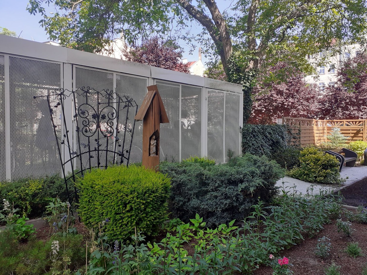 Ogród z metalową pergolą, drewnianą kapliczką i różnorodną roślinnością na tle ażurowego budynku i czerwonych drzew. Elementy małej architektury ogrodowej.