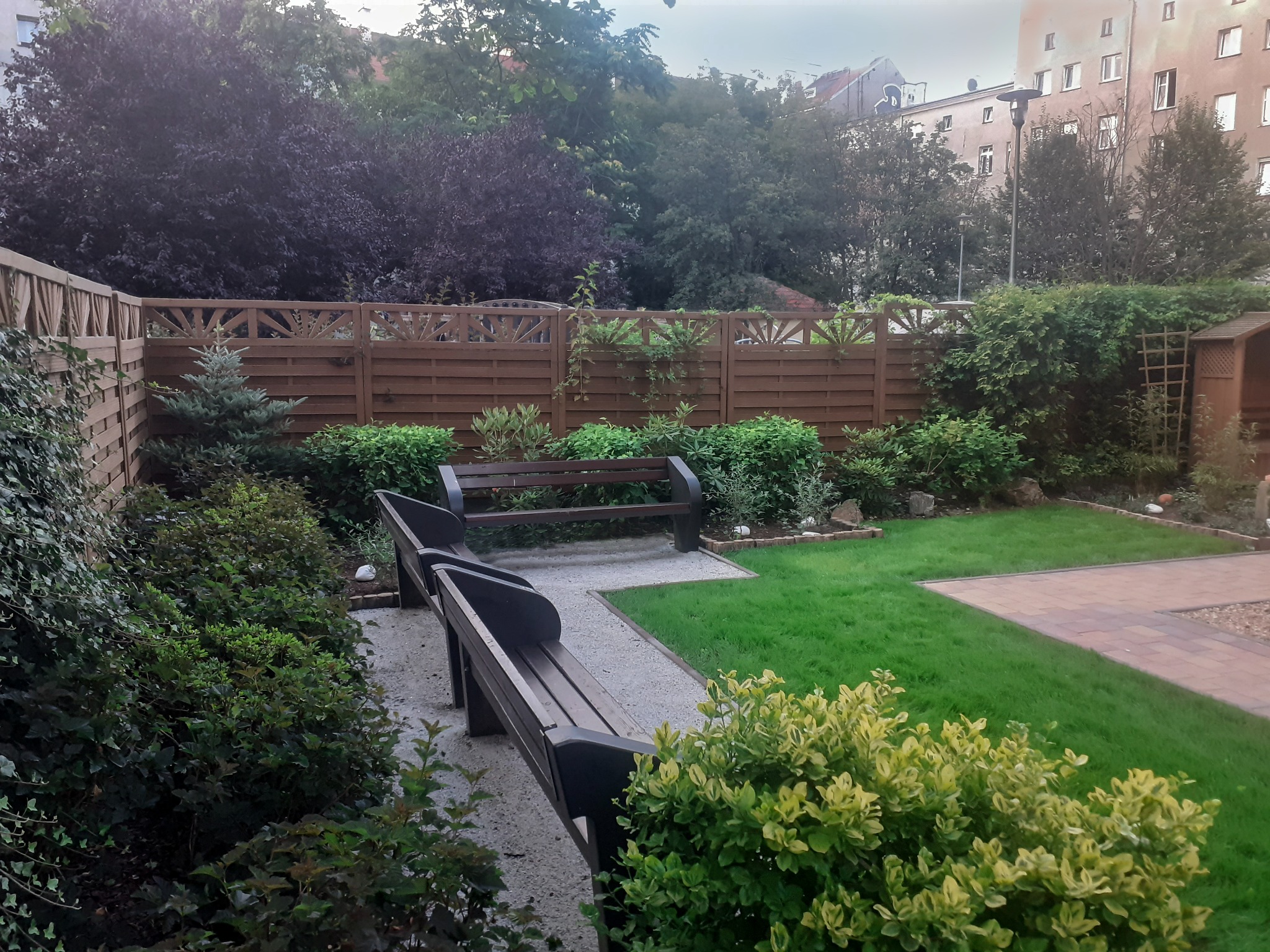 Uporządkowany ogród z dwiema ławkami, trawnikiem i ozdobnymi krzewami. Drewniany płot z ażurowym wykończeniem w tle. Elementy małej architektury ogrodowej.