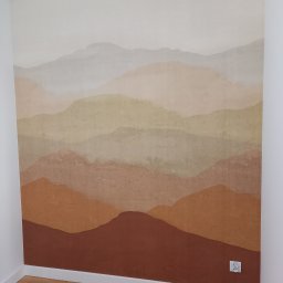 Katsiaryna Padasinavik - Ściana z ręcznie malowanym motywem górskim w odcieniach beżu, brązu i szarości, widoczna biała listwa przypodłogowa i drewniana podłoga