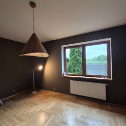 Katsiaryna Padasinavik - Pokój po malowaniu ścian na ciemny kolor, z drewnianą podłogą w jodełkę i nowoczesnym oświetleniem. Widoczne okno z widokiem na zieleń i grzejnik.