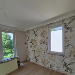 Pokój po tapetowaniu ścian z wzorem kwiatowym. Widoczne okna i drewniana podłoga. Częściowo widoczne materiały budowlane.