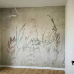 Pokój po tapetowaniu z motywem roślinnym na ścianie. Widoczne drewniane panele podłogowe, okno z widokiem na zieleń i fragment otwartych drzwi.