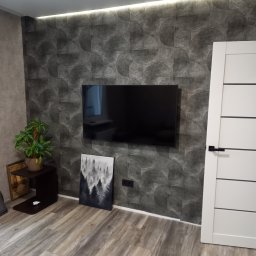 Nowoczesny salon z tapetą w geometryczne wzory, telewizorem na ścianie i drewnianą podłogą. Minimalistyczny wystrój z rośliną i obrazem leśnym.