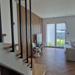 Nowoczesne wnętrze domu z drewnianą ścianą z lameli, schodami z drewnianymi stopniami i parkietem ułożonym w jodełkę. Widok na salon z oknem i sofą.