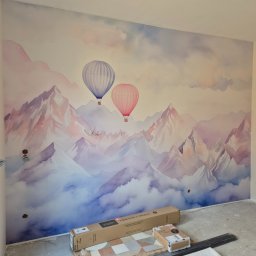 Katsiaryna Padasinavik - Ściana z tapetą w pastelowe góry i balony, kartony z panelami podłogowymi u dołu. Widoczne gniazdka elektryczne. Ukończone tapetowanie pokoju.