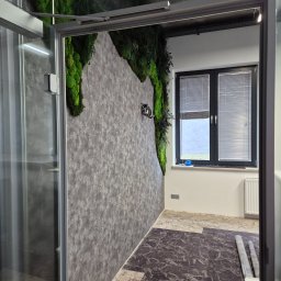 Katsiaryna Padasinavik - Nowoczesna łazienka z akcentem mchu na ścianie i wzorzystą podłogą. Widok przez otwarte drzwi kabiny prysznicowej. Wykończenie w szarościach i bieli, rolety w oknie.