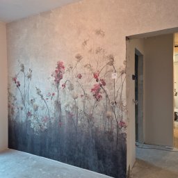 Katsiaryna Padasinavik - Ściana z fototapetą w kwiaty w odcieniach szarości i różu, widoczne gniazdka elektryczne i surowa podłoga. Ujęcie z niskiego kąta, widok na otwarte drzwi do innego pomieszczenia.