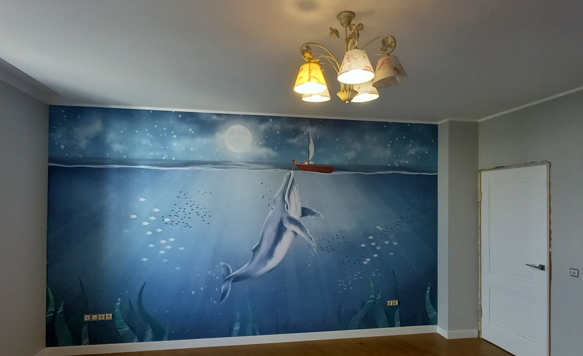Ściana pokoju dziecięcego z ręcznie malowanym muralem przedstawiającym podwodny świat z wielorybem, łodzią pod żaglami i ławicą ryb, oświetlona ozdobnym żyrandolem.