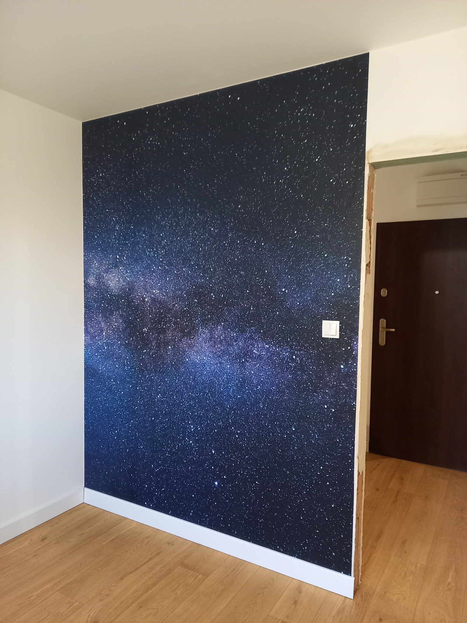 Ściana z tapetą przedstawiającą galaktykę, listwa przypodłogowa, widok na drzwi wejściowe, parkiet z paneli.
