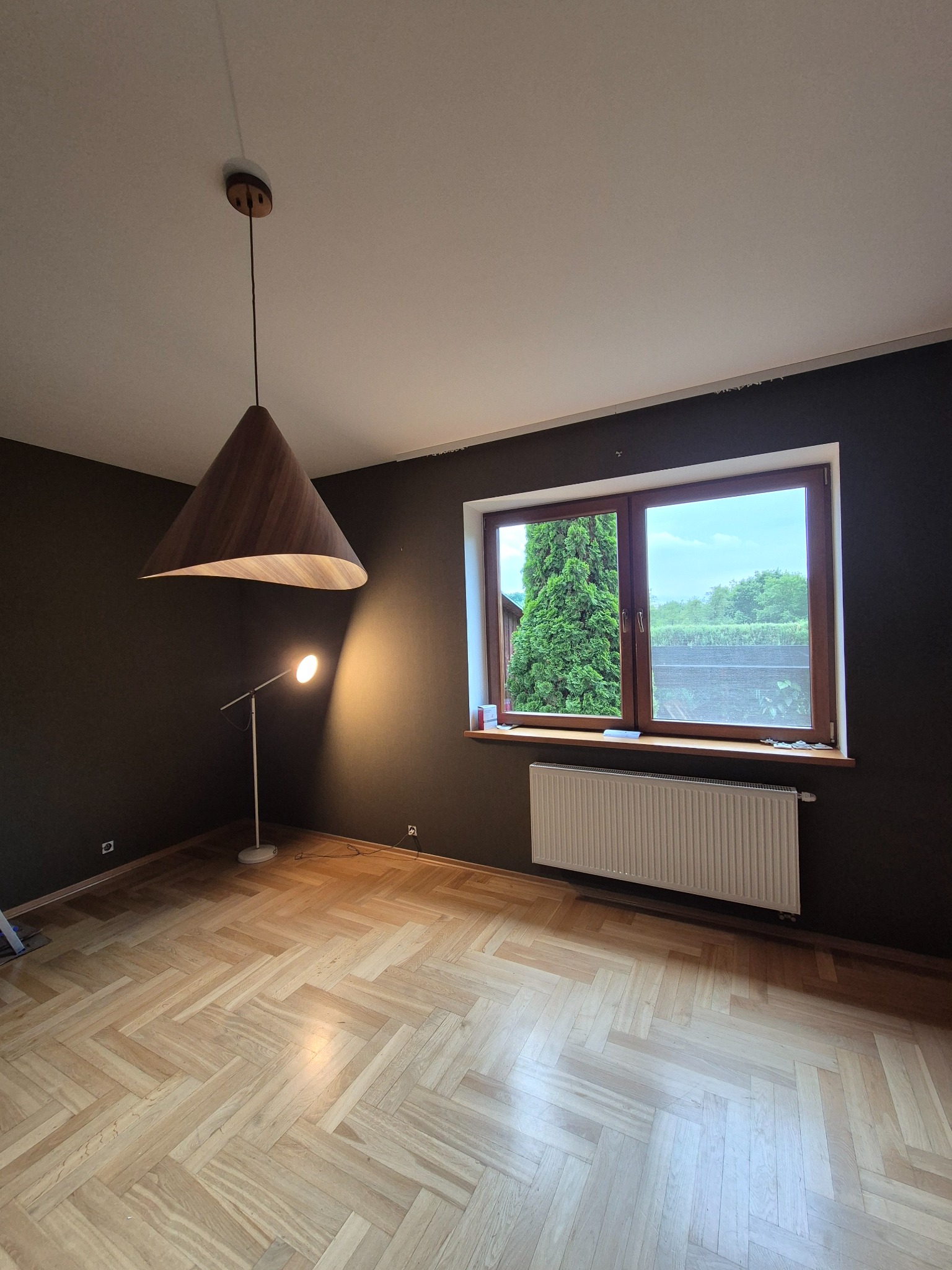 Pokój po malowaniu ścian na ciemny kolor, z drewnianą podłogą w jodełkę i nowoczesnym oświetleniem. Widoczne okno z widokiem na zieleń i grzejnik.