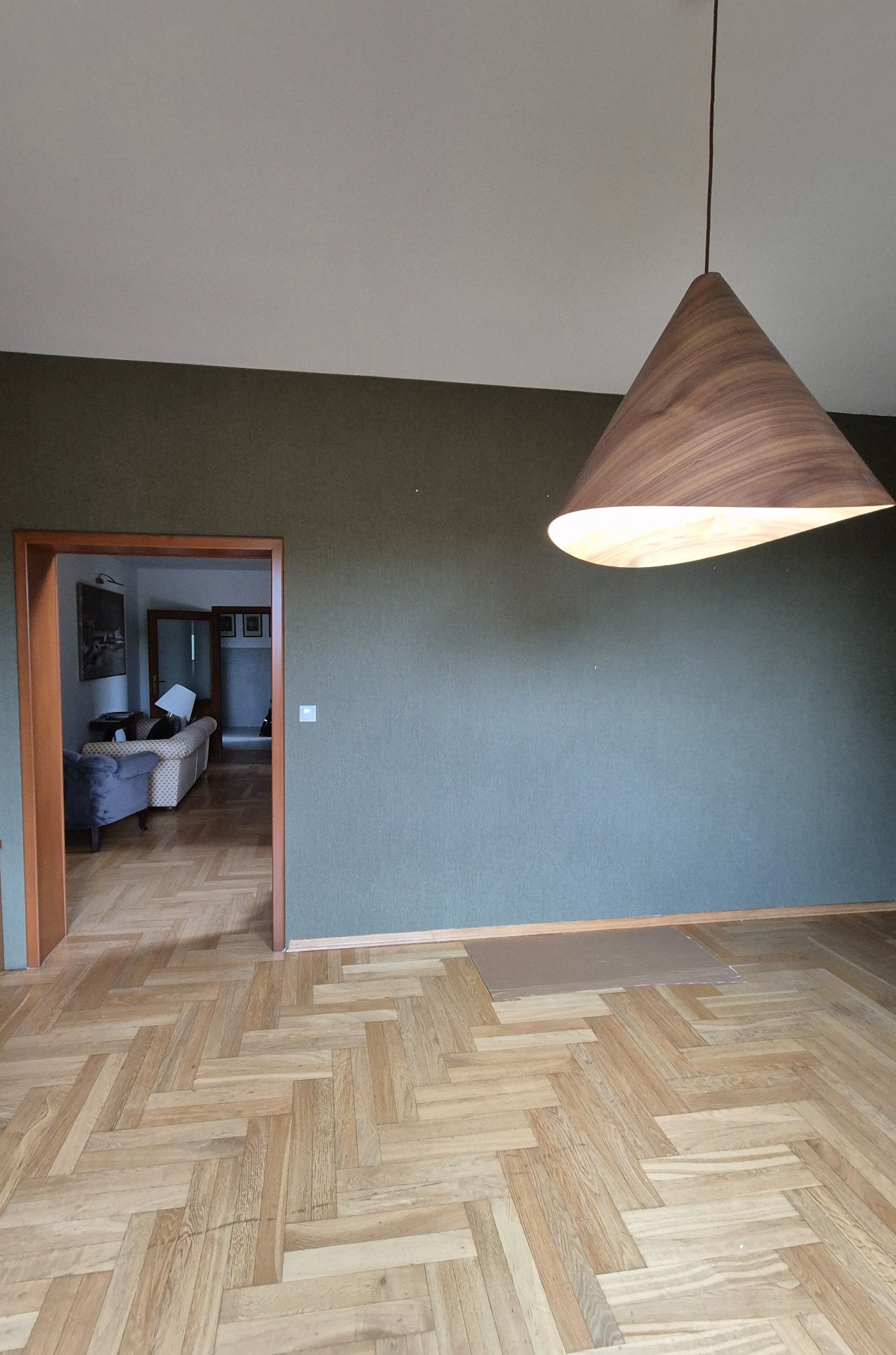 Pokój po remoncie: parkiet w jodełkę, zielona ściana z fakturą, drewniana lampa stożkowa. Widok na salon przez otwarte drzwi. Widoczne ślady prac wykończeniowych.