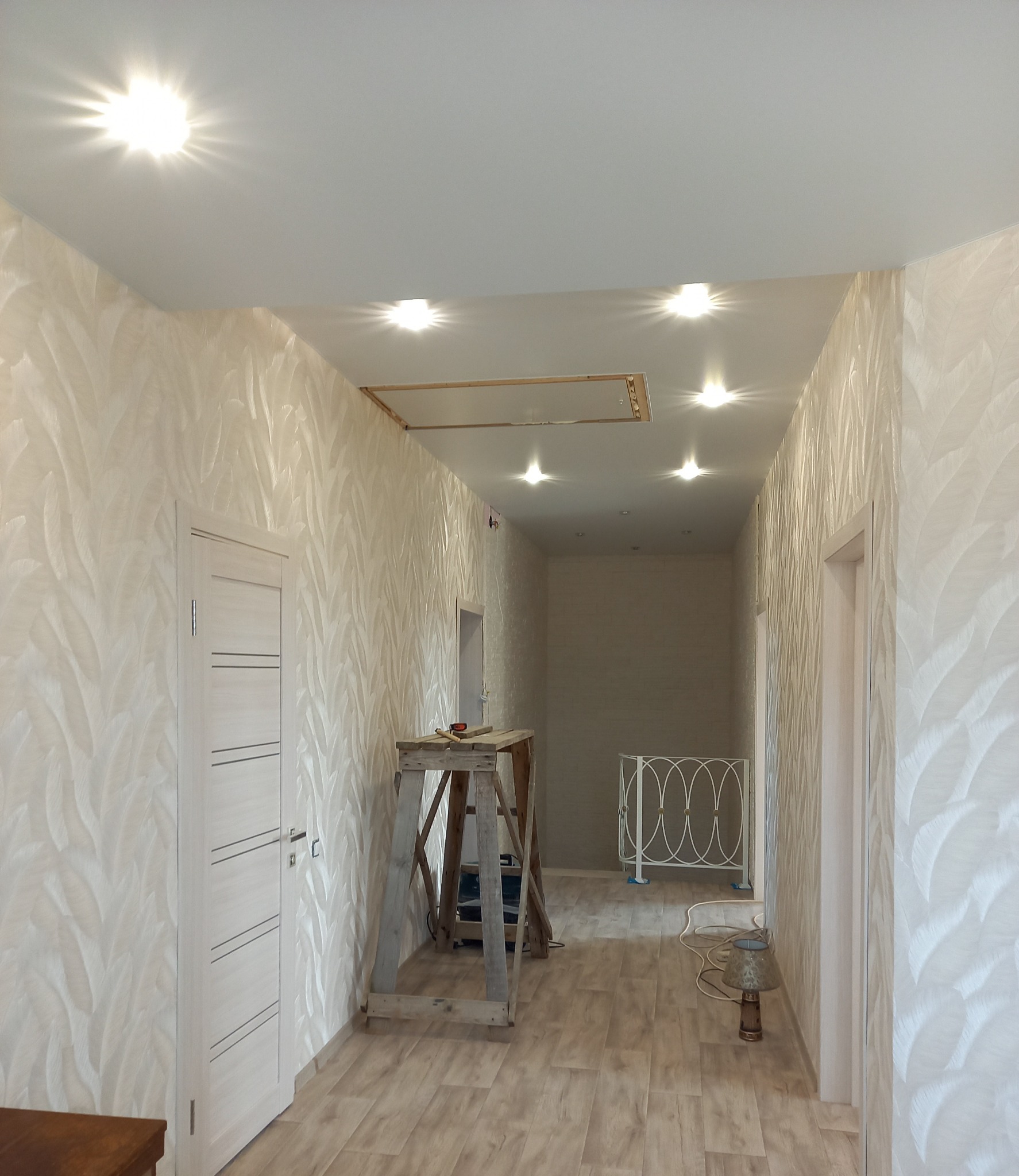 Korytarz po remoncie: wzorzyste tapety w odcieniach beżu, drewniana drabina, oświetlenie sufitowe. Widoczny efekt tapetowania i częściowo wykończone wnętrze.