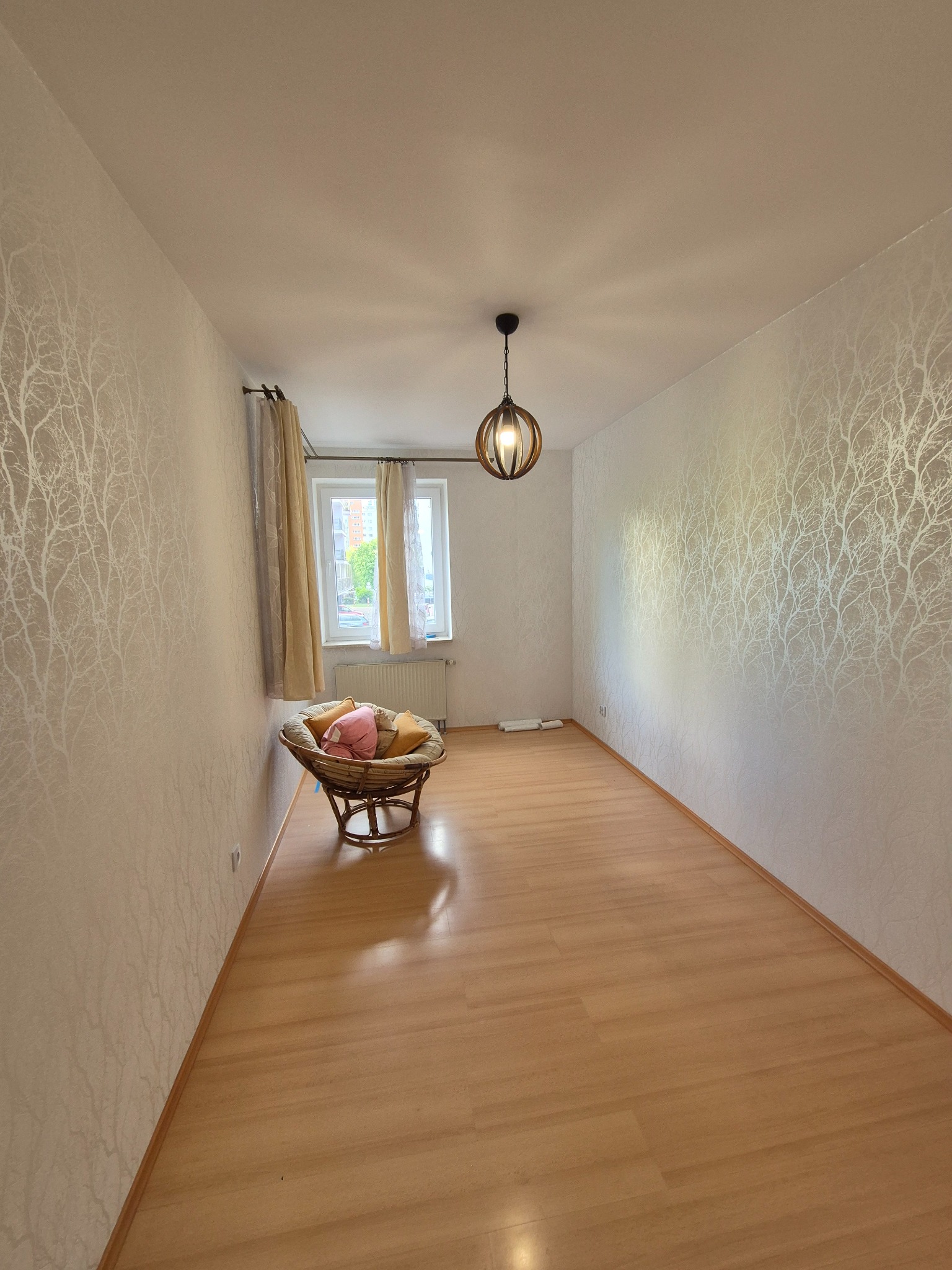 Pokój po tapetowaniu z motywem drzew, z drewnianą podłogą i wiklinowym fotelem. Widoczne okno z zasłonami i lampa sufitowa.