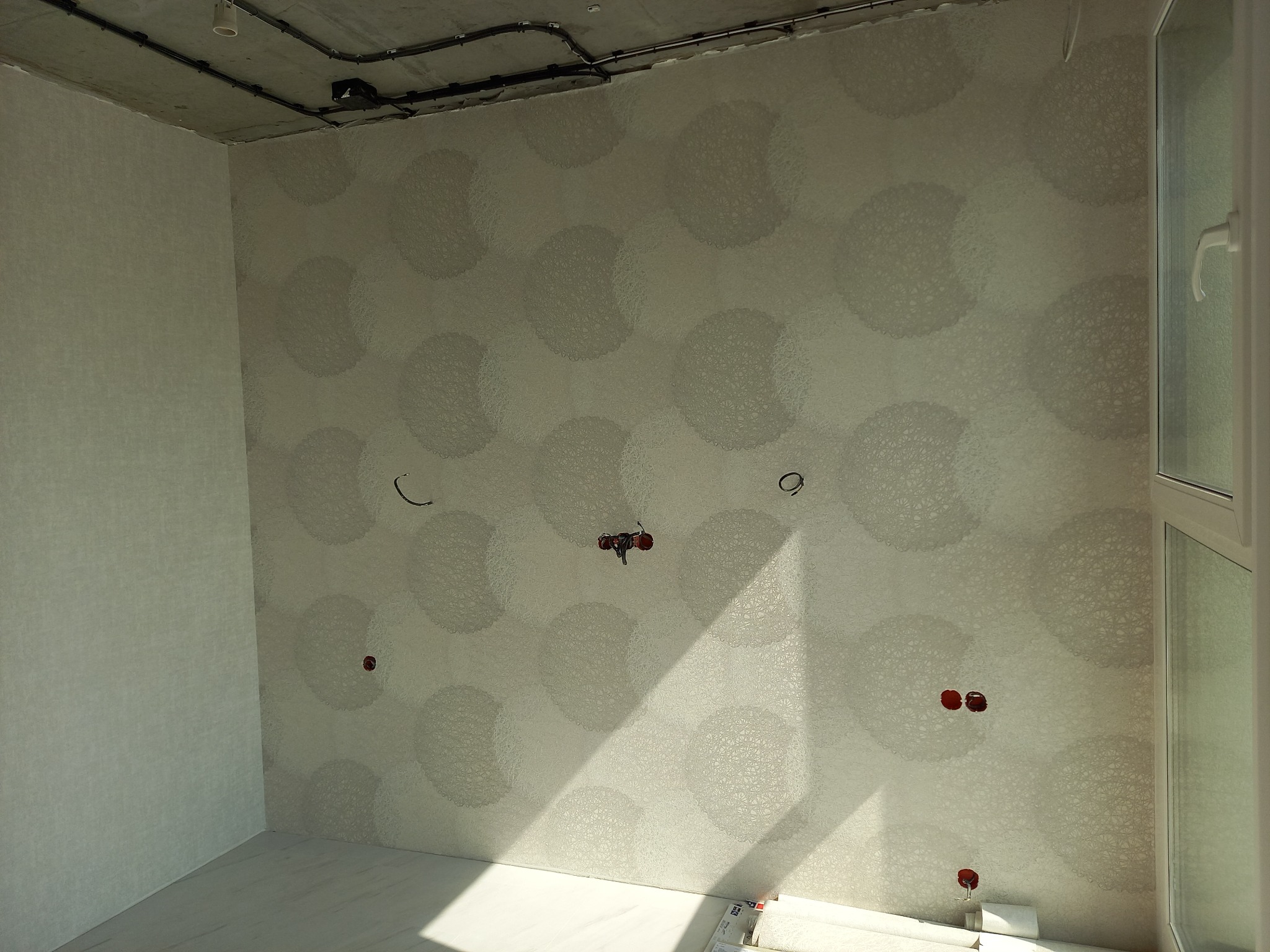 Pokój po tapetowaniu: wzorzysta tapeta w jasnych odcieniach na ścianie, widoczne gniazdka elektryczne, surowy betonowy sufit z instalacją.